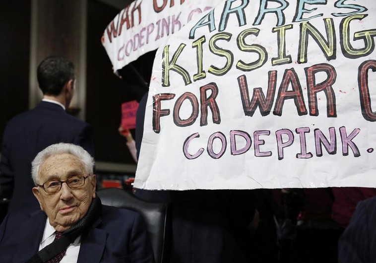 Former United States Secretary of State Henry Kissinger sits calmly as protesters demand his arrest