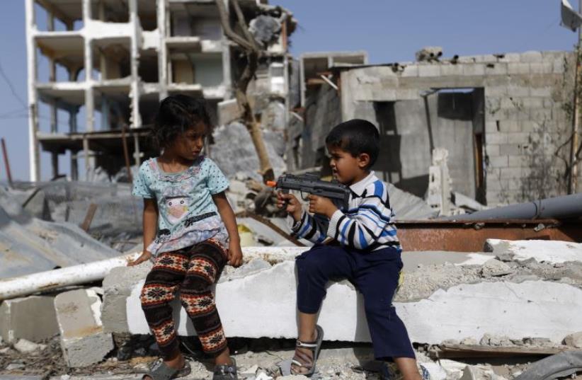Palestinian children play with a toy gun in front of homes ruined during Israel's bombardment in 2014 (photo credit: MOHAMMED ABED / AFP)