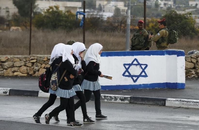PALESTINIAN SCHOOL girls walk past Israeli soldiers at a checkpoint at a entrance to the village of Beit Einun near Hebron (photo credit: REUTERS)