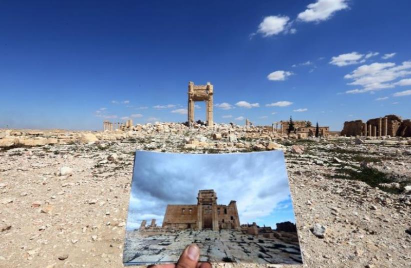 A general view taken on March 31, 2016 shows a photographer holding his picture of the Temple of Bel taken on March 14, 2014 in front of the remains of the historic temple after it was destroyed by Islamic State (photo credit: AFP PHOTO)