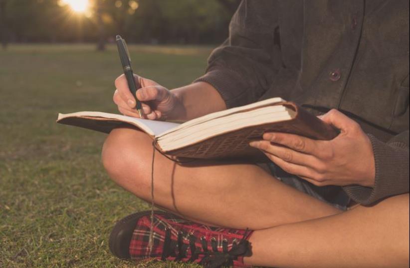 A young woman is sitting on the grass in a park at sunset and is writing in a notebook (illustrative) (photo credit: INGIMAGE)
