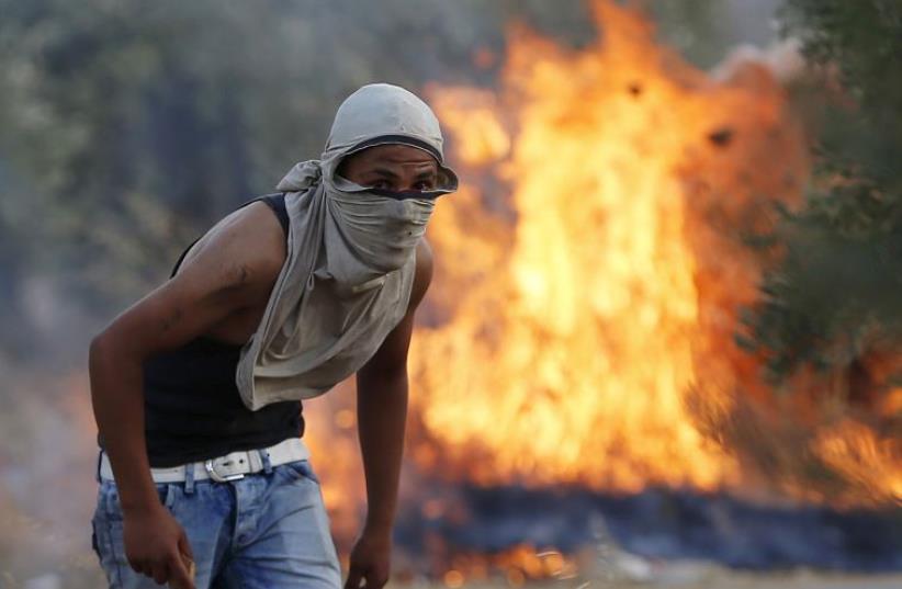 A Palestinian stone-thrower looks on as he stands in front of a fire during clashes with IDF troops in the West Bank village of Duma (photo credit: REUTERS)