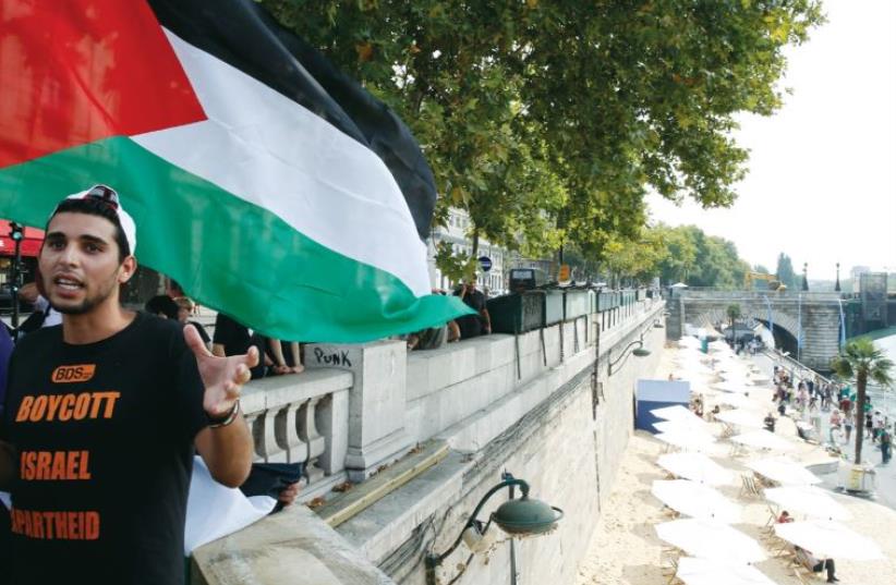 A man wearing a T-shirt that says, ‘Boycott Israel Apartheid’ holds a Palestinian flag during a protest, while below, an artificial beach is set up with the theme ‘Tel Aviv on the Seine,' (photo credit: REUTERS)
