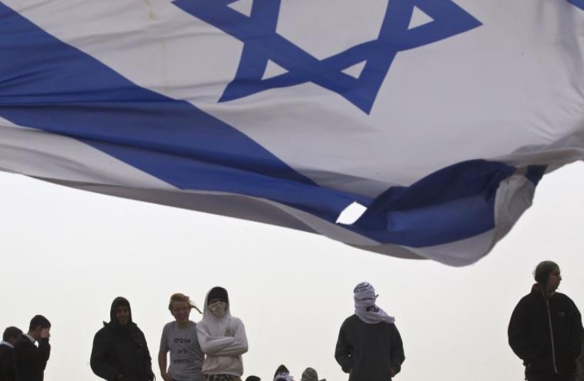 An Israeli flag flutters near Israeli youths as they protest atop the roof of a synagogue in the Israeli settlement of Givat Ze'ev (photo credit: REUTERS)