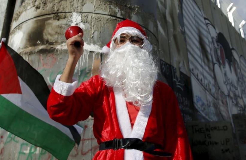 A Palestinian protester wearing Santa Claus costume stands in front of a section of the Israeli barrier during an anti-Israel protest in the West Bank city of Bethlehem December 18, 2015 (photo credit: REUTERS)