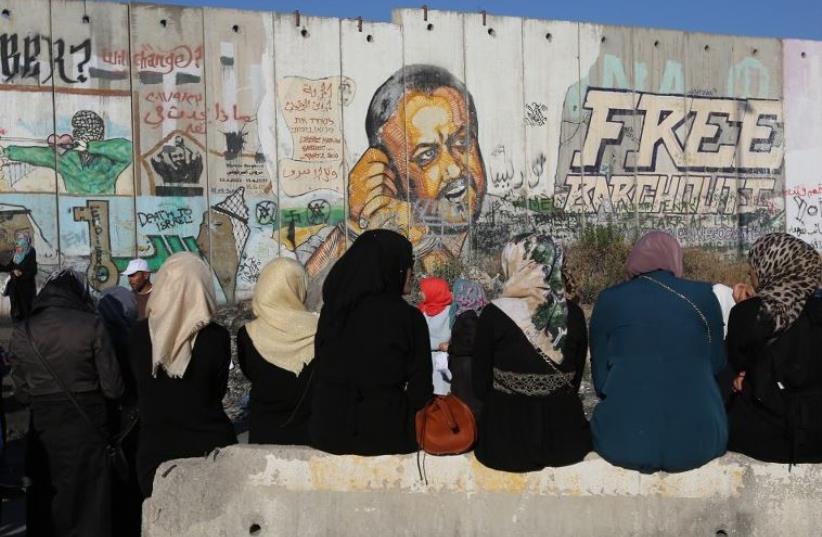 Palestinian women sit near a section of Israel's separation barrier at Qalandia checkpoint in front of a painted mural of Marwan Barghouti (photo credit: AFP PHOTO)
