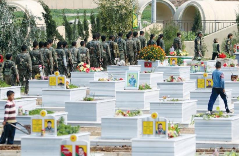 KURDISH PEOPLE’S Protection Units (YPG) fighters walk near graves of fellow fighters during a visit to a YPG graveyard in Qamishli, Syria, on March 11. (photo credit: REUTERS)