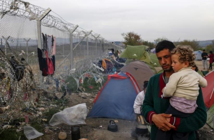 A man with a child looks at the border fence at a makeshift camp for migrants and refugees at the Greek-Macedonian border (photo credit: REUTERS)