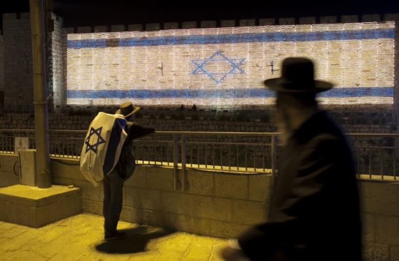 A man with an Israeli flag covering his backpack looks at an Israeli flag made of lights installed on the wall of Jerusalem's Old City (photo credit: REUTERS)
