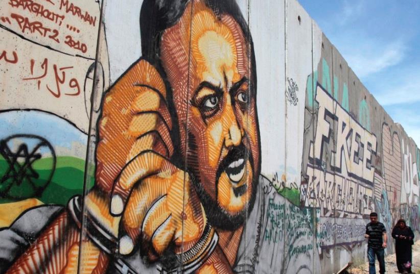PALESTINIANS WALK past graffiti depicting jailed Fatah leader Marwan Barghouti, currently serving five life terms for terrorism, on a section of the security wall on the road to Ramallah. (photo credit: REUTERS)