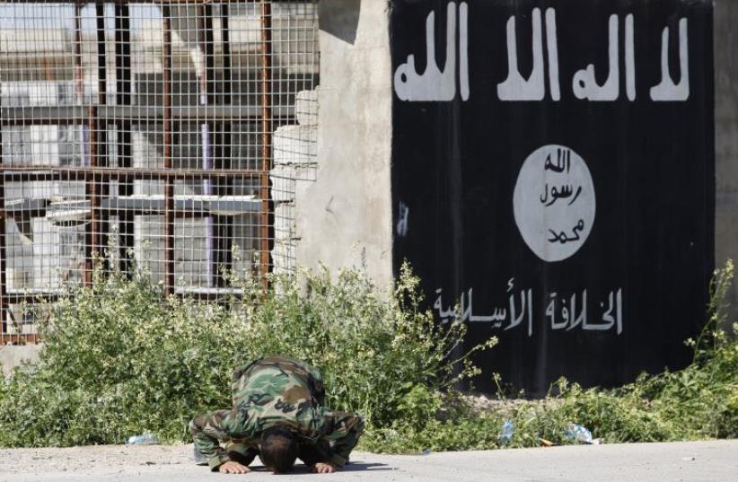 A member of a militia kneels as he celebrates victory next to a wall painted with the black flag commonly used by ISIS militants (photo credit: REUTERS)